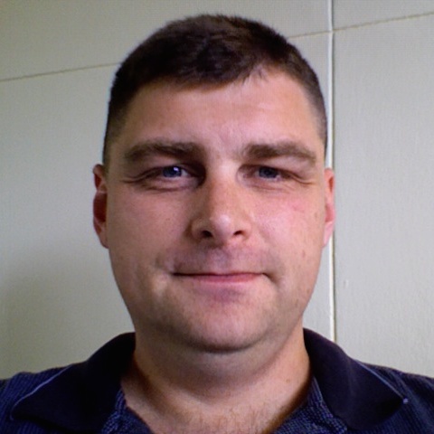 Portrait photograph of Erik, a clean-shaven white man with short, dark hair wearing a navy blue polo shirt on a white background. He is smiling slightly